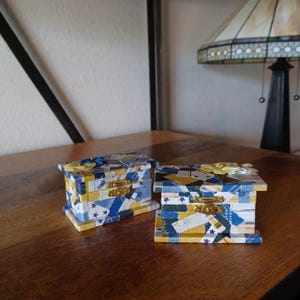 May include: Two small decorative boxes with a patchwork design in shades of blue, yellow, and white. The boxes have gold hinges and clasps.