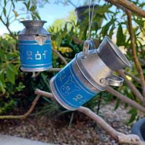 May include: Two miniature metal milk cans with blue bands and Japanese text hang from a tree branch. The cans have a silver finish and a handle. One can features a cow graphic.