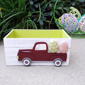 May include: A white wooden box with a green lid. The box has a red truck painted on the front with three pastel Easter eggs in the truck bed.
