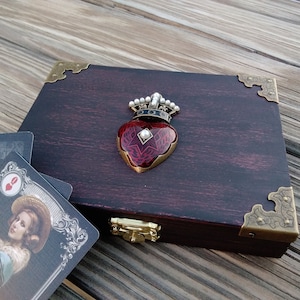 May include: A dark wood box with brass corners and a hinged lid. The box has a red heart with a crown and a pearl on top. The box is sitting on a wooden surface.