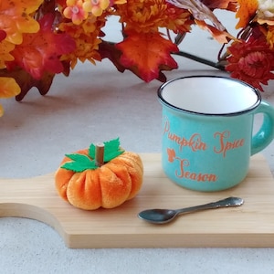 May include: A teal mug with the text "Pumpkin Spice Season" in orange lettering, a small orange velvet pumpkin, and a silver teaspoon on a wooden chopping board.