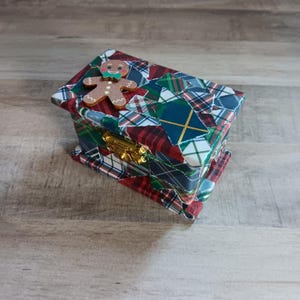 May include: A small wooden box with a plaid pattern in red, green, blue, and white. The box has a gingerbread man figure on the top and a gold latch.