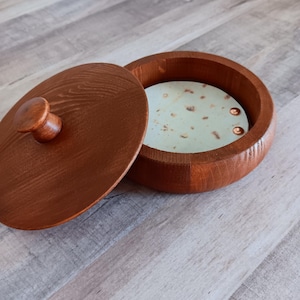 May include: A round, wooden box with a lid. The box and lid are a warm brown color. The lid is slightly ajar, revealing a light green interior with copper-colored accents. The box sits on a light gray wooden surface.