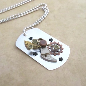 May include: A silver dog tag necklace with a steampunk design. The tag features a variety of gears, cogs, and other metal pieces, all arranged in a whimsical pattern. The necklace is made of a silver chain.