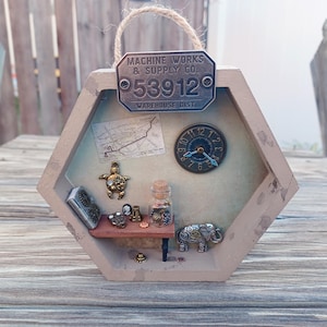 May include: A steampunk-themed shadow box with a hexagonal frame. It features miniature metal objects, including a clock, map, turtle, elephant, and a small book. The top has a metal sign that reads "MACHINE WORKS & SUPPLY CO. 53912 WAREHOUSE DIST." A jute rope is attached for hanging.