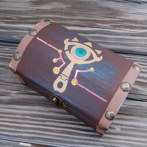 May include: A wooden treasure chest with a brown, pink, and blue paint finish. The lid features a gold-colored Sheikah Eye symbol with a blue eye and a gold-colored handle.