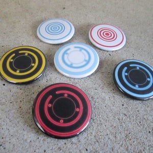May include: Six round buttons with different colored and patterned designs. The buttons feature a black center circle with concentric circles around it. The colors of the circles are red, blue, yellow, and black.