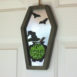 May include: A wooden coffin-shaped mirror with a green cauldron, a witch's hat, and bats cut out of the mirror. The mirror has a silver backing.