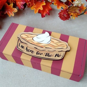 May include: A rectangular wooden box with burgundy and yellow stripes. A wooden pie-shaped decoration with whipped cream and the text "I'm here for the pie" is on top. Autumn leaves are in the background.