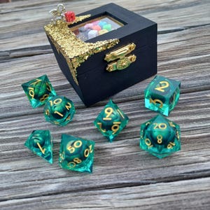 May include: A black wooden box with a gold glitter trim and a clear window, containing colorful dice. Several green and gold dice are scattered around the box, showcasing various shapes and numbers. A decorative piece sits atop the box.