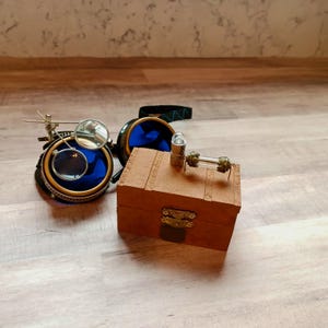 May include: A pair of steampunk goggles with blue lenses and gold accents, a small wooden box with a brass latch, and a glass tube with a metal cap.
