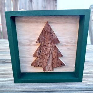 May include: A wooden Christmas tree decoration. The tree is made of textured bark and is set against a light wood background. The frame is a dark green color.