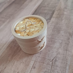 May include: A small, cylindrical wooden container with a woven bamboo lid and a twine handle. The container is light beige with decorative twine accents. The lid is made of woven bamboo strips. The container is sitting on a wooden surface.