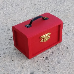 May include: A small red wooden box with a black handle and a gold latch. The box has a rounded top and a rectangular base.