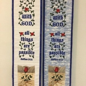 May include: Two white and blue fabric wall hangings with the text "With God all things are possible" and "Matthew 19:26" embroidered on them. The hangings feature a red rose with green leaves and stems.