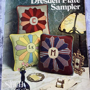 May include: A Leisure Arts Dresden Plate Sampler leaflet 272 featuring three square pillows with circular patchwork designs and monograms C, LK, and M. The cover includes a lamp, a blue vase with flowers, a silver tray, and white gloves.