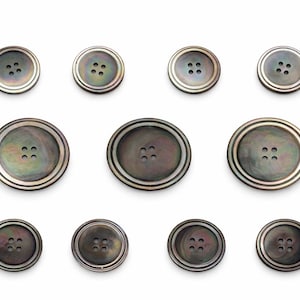 May include: A collection of dark gray buttons with a pearlescent sheen. The buttons vary in size and feature four holes each. Each button has a double-layered, silver-colored rim. The buttons are arranged on a white background.