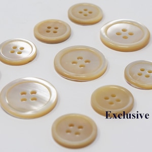 Może przedstawiać: Kolekcja beżowych guzików w różnych rozmiarach, każdy z czterema otworami. Guziki mają perłowy połysk i są ułożone na białej powierzchni. Widoczny jest tekst "Exclusive Buttons".