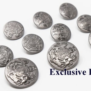 Puede incluir: Diez botones plateados con un diseño en relieve que presenta un escudo de armas. Los botones están dispuestos en dos filas sobre una superficie blanca. El texto "Exclusive Buttons" es visible en la parte inferior de la imagen.