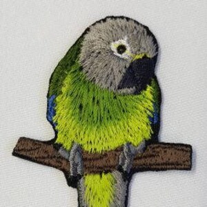 May include: Embroidered iron-on patch of a green and gray parrot perched on a brown branch. The parrot has a yellow chest and a blue patch on its wing.