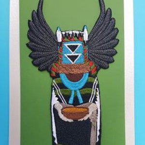 Könnte beinhalten: Gestickter Patch einer amerikanischen Kachina-Puppe mit schwarzen Flügeln, einem blau-grünen gemusterten Kleid und einer Schale in den Händen. Der Patch befindet sich auf einem grünen Hintergrund mit einem schwarzen Etikett, auf dem "Tangled Threads Embroidery" steht.