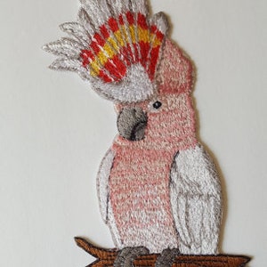 May include: Embroidered iron-on patch of a pink and white cockatoo perched on a branch. The cockatoo has a red and yellow crest.