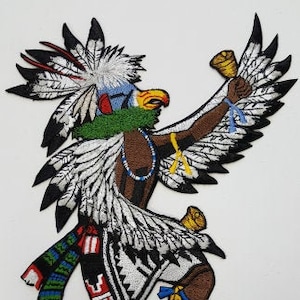 May include: Embroidered patch of a Native American dancer wearing a feathered headdress and holding a bell. The dancer is wearing a colorful outfit with geometric patterns and bells on their ankles.