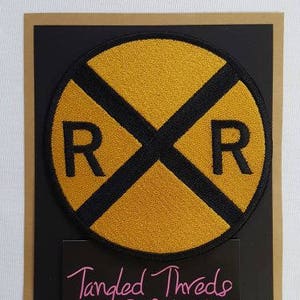 May include: A yellow and black embroidered iron-on patch with the letters "RR" in a circle. The patch is for sewing or ironing onto clothing, bags, or other fabric items. The text "Tangled Threads Embroidery" is visible below the patch.