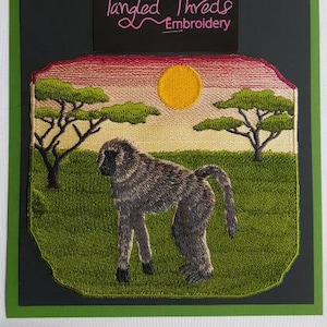 May include: Embroidered patch depicting a baboon in a savanna scene with a sunset sky. The patch features a pink, orange, and yellow sunset with green trees and grass. The baboon is brown and grey with a long tail. The patch is bordered in black and includes the text "Tangled Threads Embroidery" in pink.