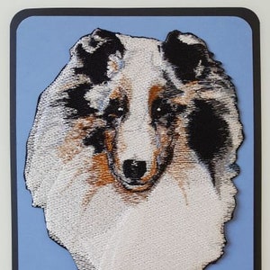 May include: Embroidered patch of a white, brown, and black dog with fluffy fur. The dog is looking directly at the viewer.