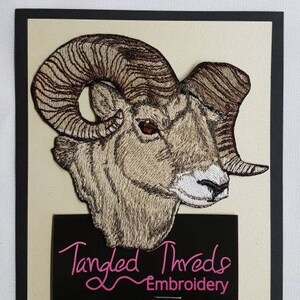 May include: Embroidered patch of a brown and white ram with large curled horns. The patch is on a beige background with a black label that reads "Tangled Threads Embroidery".