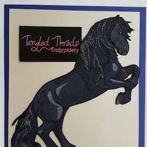 May include: An embroidered image of a rearing black horse with a flowing mane and tail. The horse is set against a cream background with a dark blue border. A black rectangle with the text "Tangled Threads Embroidery" is in the upper left corner.