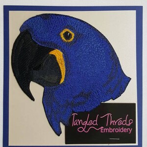 May include: Embroidered patch of a blue hyacinth macaw's head with a black beak and yellow accents. The patch is on a beige background with a black tag that reads "Tangled Threads Embroidery".