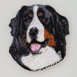 May include: Embroidered patch of a Bernese Mountain Dog with black, white, and brown fur. The dog is looking directly at the viewer with its tongue sticking out.