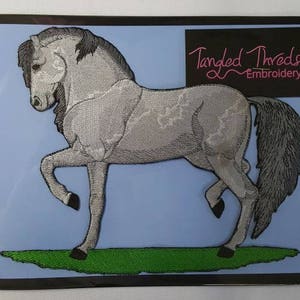 Könnte beinhalten: Ein gestickter Pferde-Patch in Grau mit schwarzer Mähne und Schweif. Der Patch befindet sich auf einem blauen Hintergrund mit grünem Gras am unteren Rand. Der Text "Tangled Threads Embroidery" befindet sich oben rechts.