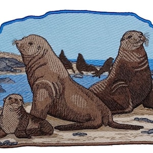 May include: Three brown sea lions on a sandy beach with a blue ocean and rocky shore in the background.