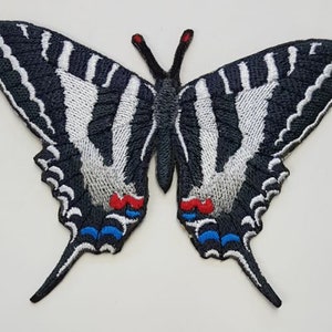 以下が含まれることがあります： 赤と青のアクセントが付いた、黒と白の刺繍された蝶のワッペン。蝶の翼を広げた長さは約7.5cmです。
