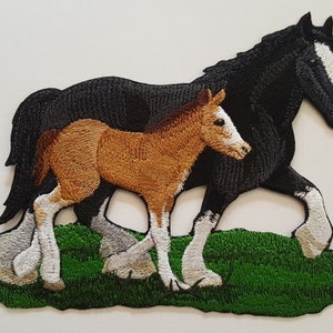 May include: Embroidered patch of a black horse and a brown foal standing in a green field. The horse has a white stripe on its face and white markings on its legs. The foal has white markings on its legs.