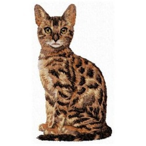 May include: A brown and black tabby cat with a spotted coat is sitting with its head tilted slightly to the side. The cat has green eyes and a white patch on its chest.