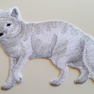 May include: White embroidered iron-on patch of an arctic fox. The fox is standing with its head turned to the left. The patch is perfect for adding a touch of nature to your clothing or crafts.