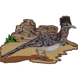 May include: Embroidered patch depicting a roadrunner bird standing on a desert landscape with brown rocks and green plants. The bird is brown, black, and white with a long tail and a pointed beak.
