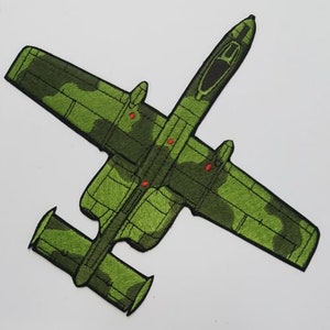 May include: A green camouflage embroidered patch of an A-10 Warthog aircraft. The patch is detailed with red stitching for the aircraft's cannons.
