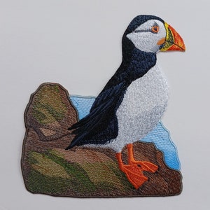 Può includere: Toppa ricamata di un puffin in piedi su uno scoglio con uno sfondo blu. Il puffin è nero e bianco con un becco arancione.
