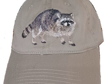 May include: A beige baseball cap with an embroidered raccoon design. The raccoon is facing forward with its head tilted slightly to the side. The raccoon is brown and gray with black eyes and a black nose.