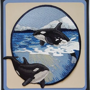 May include: Embroidered patch featuring two black and white orcas swimming in a blue ocean with white waves. The orcas are jumping out of the water with their tails in the air. The background is a light blue sky with white clouds.