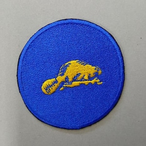 Puede incluir: Un parche bordado azul con un contorno amarillo de un castor.