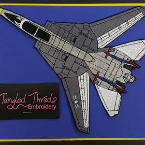 Puede incluir: Parche bordado de un avión de combate gris y blanco con detalles rojos y azules. El parche está sobre un fondo azul y tiene el texto "Tangled Threads Embroidery" en rosa.