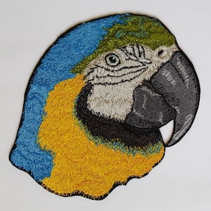 May include: Embroidered patch of a blue and yellow macaw parrot's head. The parrot has a black beak and green feathers on its head.
