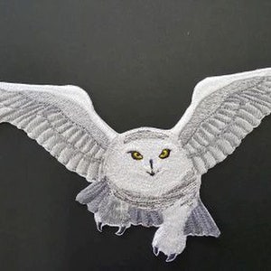 May include: White snowy owl with yellow eyes, wings spread wide, flying in mid-air. The owl is embroidered on a white fabric patch.