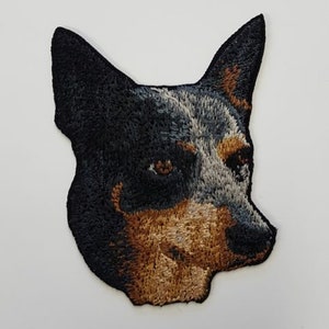 May include: Embroidered patch of a blue heeler dog with black and brown fur. The dog is looking to the left with its tongue out.
